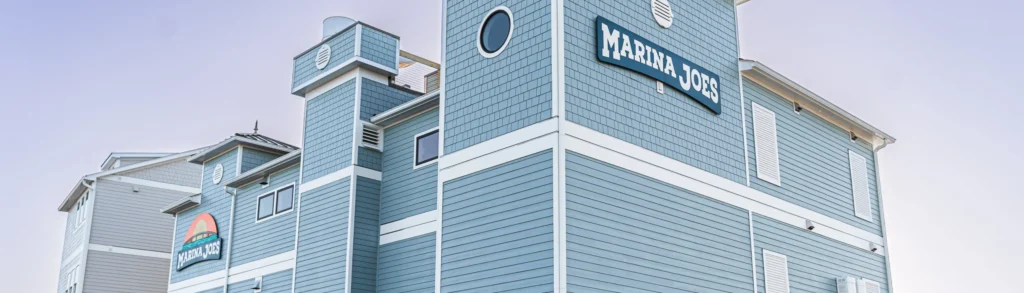 Exterior view of the Marina Joe's multi-story light blue building with branded signage and parking lot at dusk, the perfect day at Marina Joe's starts here.