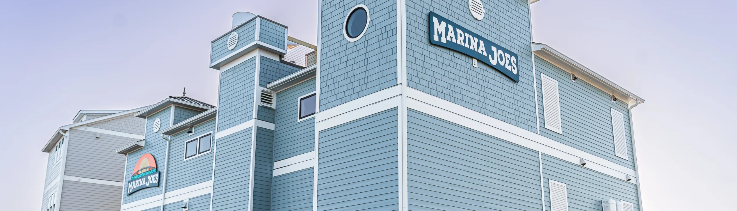 Exterior view of the Marina Joe's multi-story light blue building with branded signage and parking lot at dusk, the perfect day at Marina Joe's starts here.