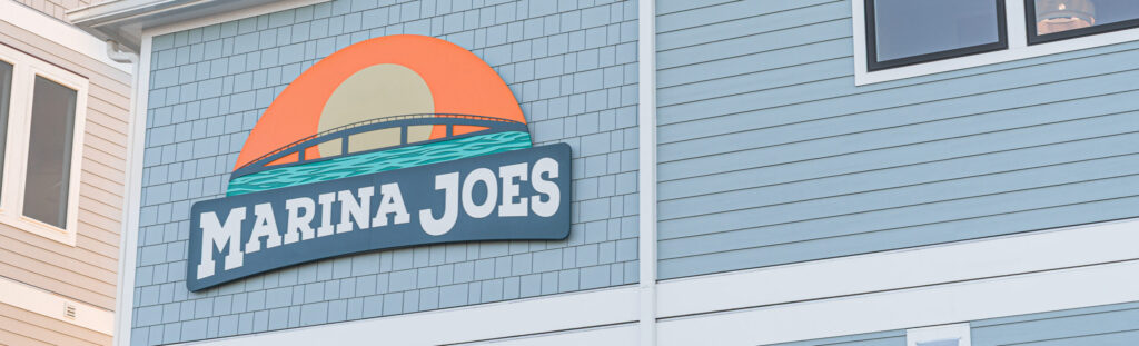 Close-up of the Marina Joe's colorful sunset and bridge logo sign on the light blue exterior building at Surf City NC, a top destination for where to take dad for Father's Day at Topsail Island
